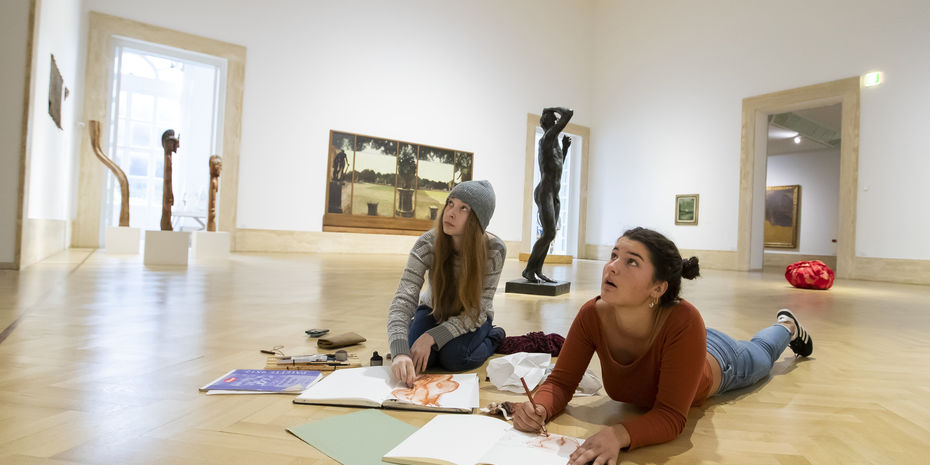 Two students drawing in their sketchbooks on the floor of a museum