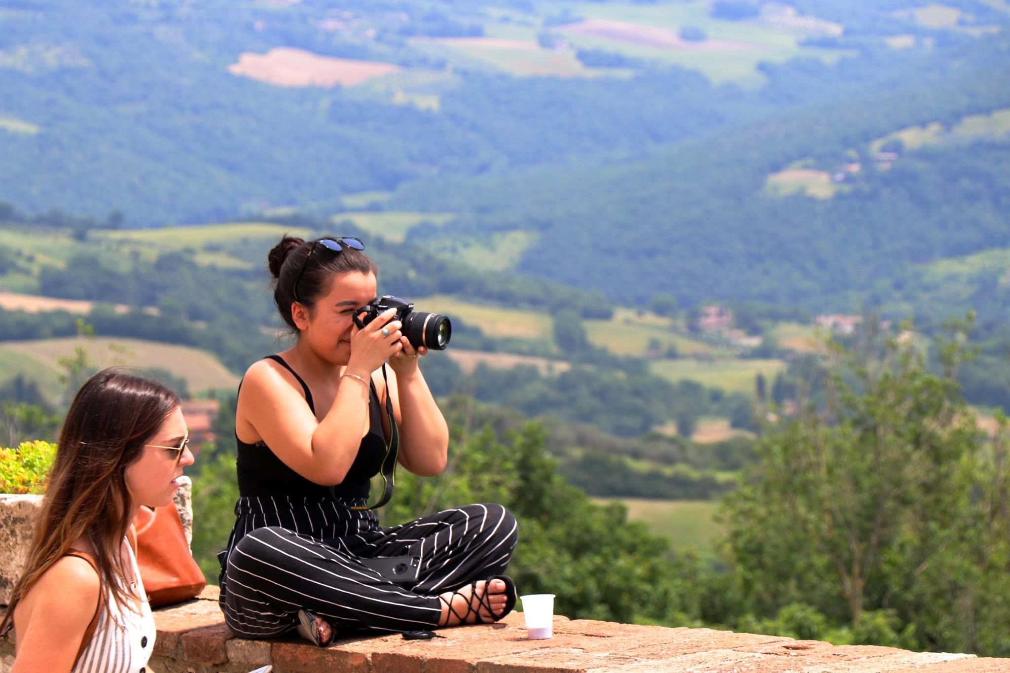 Female student sitting on ledge taking a photo, with another female student to her left. Italian countryside scenery in background.