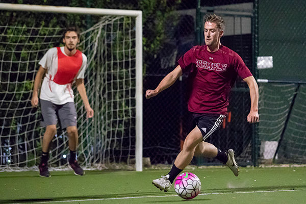 Male student in Temple t-shirt playing soccer, with male student in red jersey as goalie in the background.