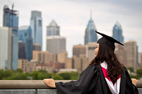 A female student in a graduation cap and gown stands in front of Philadelphia skyline
